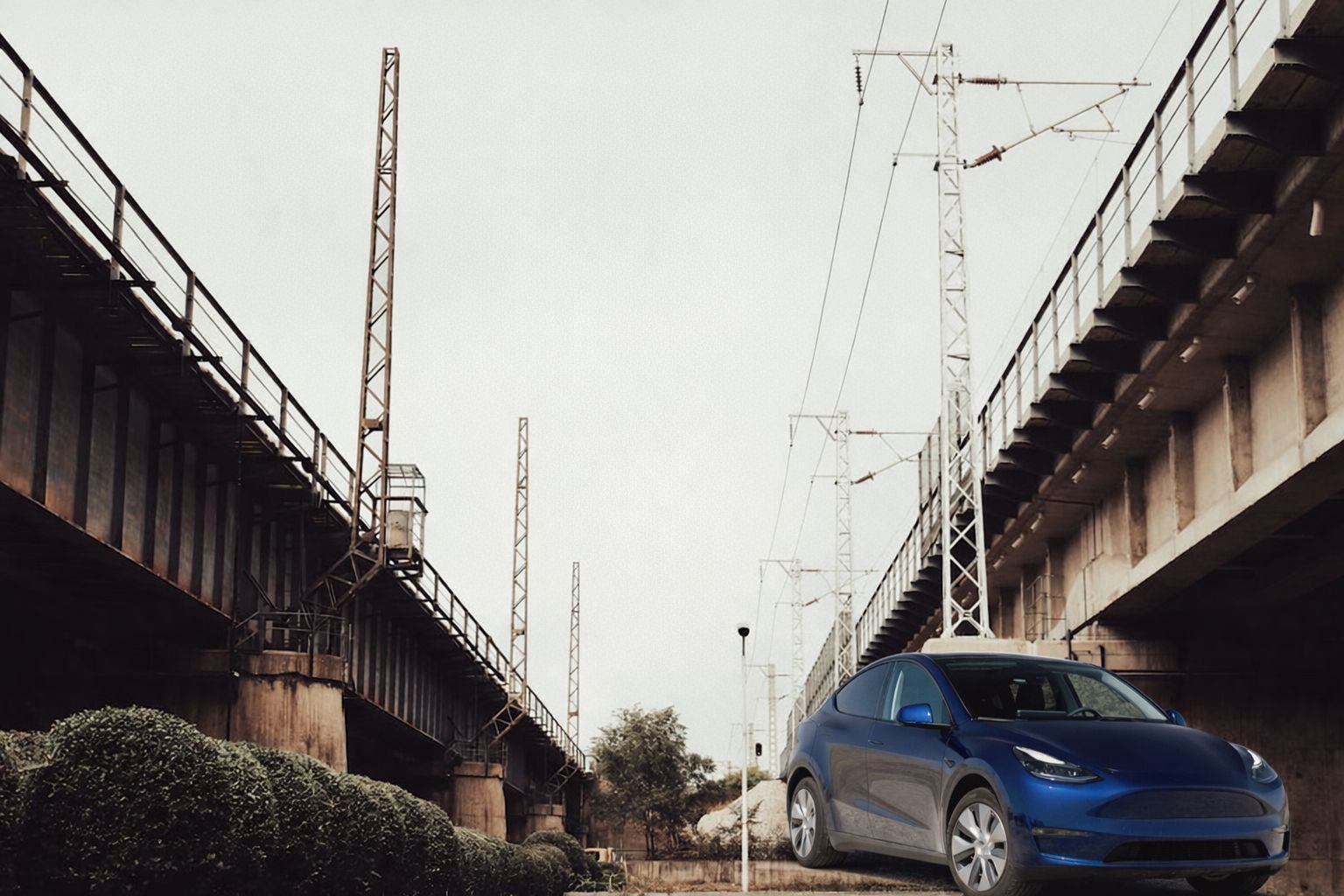 Sähköauto parkissa rautatiesiltojen ja sähkölinjojen alla, symboloi infrastruktuuria.